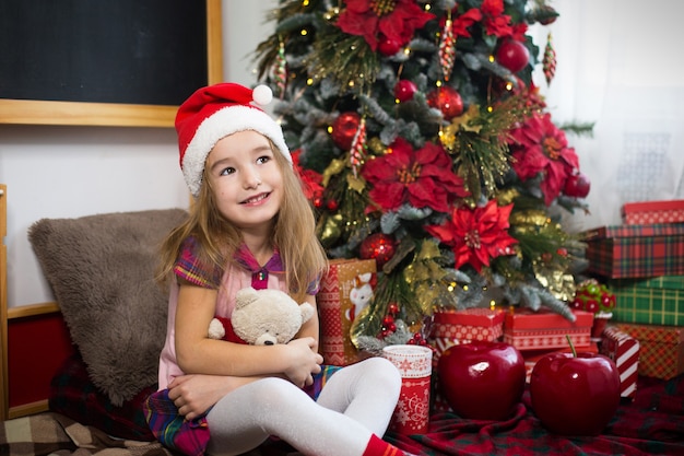 A little girl holding a Teddy bear, sitting on a plaid blanket in the Christmas decorations near a Christmas tree with boxes of gifts and a Santa hat. New year, children's game