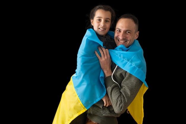 우크라이나 국기를 들고 어두운 배경에서 아빠를 껴안고 있는 어린 소녀가 우크라이나와 함께 서 있습니다.