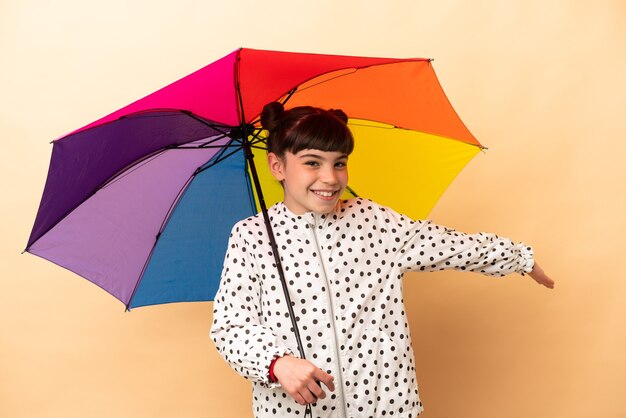 Little girl holding an umbrella isolated on beige extending hands to the side for inviting to come