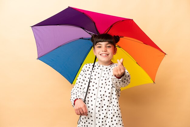 Bambina che tiene un ombrello isolato su beige che fa gesto di soldi