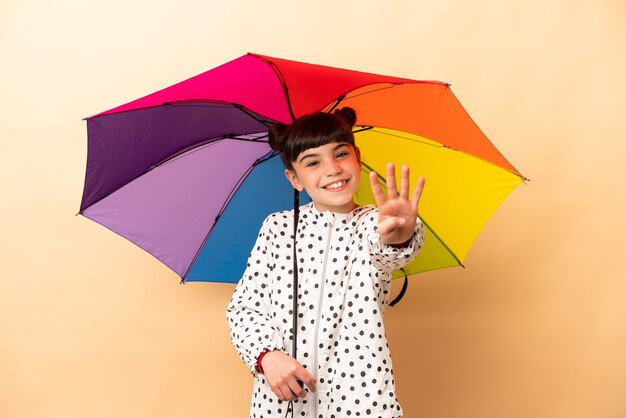 Little girl holding an umbrella isolated on beige wall happy and counting four with fingers