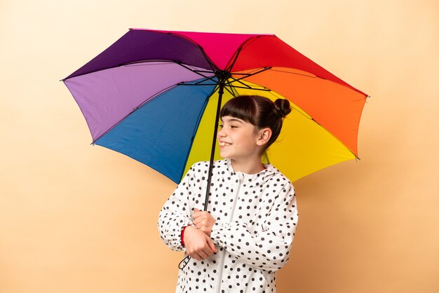 Little girl holding an umbrella isolated on beige wall looking side