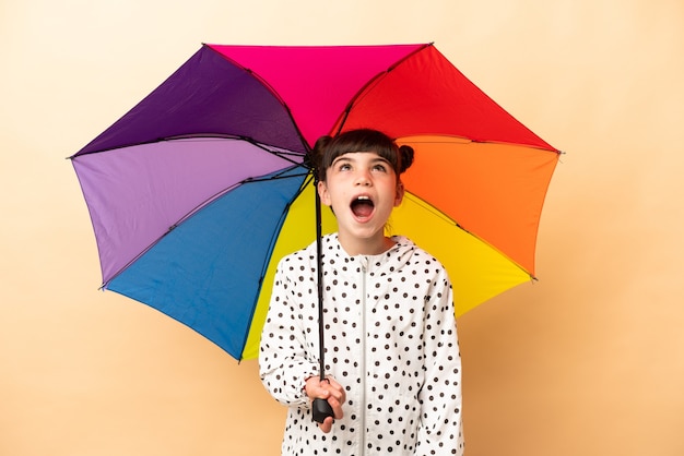 Little girl holding an umbrella isolated on beige wall looking up and with surprised expression