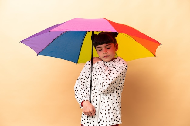 노력을 한 데 대한 어깨에 통증을 앓고 베이지 색 벽에 고립 된 우산을 들고 어린 소녀