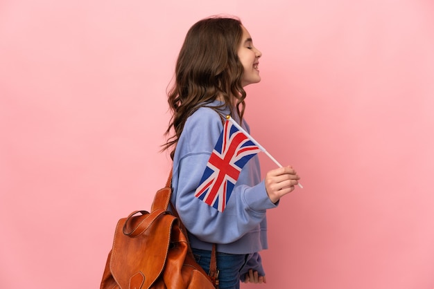 Little girl holding an United Kingdom flag isolated on pink background laughing in lateral position