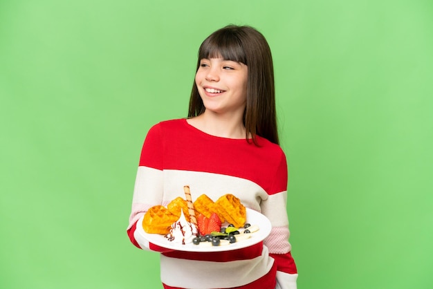 Little girl holding waffles over isolated chroma key background looking to the side and smiling