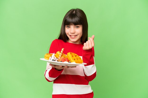 Little girl holding waffles over isolated chroma key background making money gesture