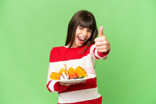 Photo little girl holding waffles over isolated chroma key background with thumbs up because something good has happened