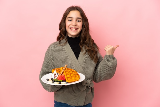 Little girl holding waffles isolated on pink wall pointing to the side to present a product