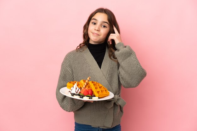 Little girl holding waffles isolated on pink wall thinking an idea