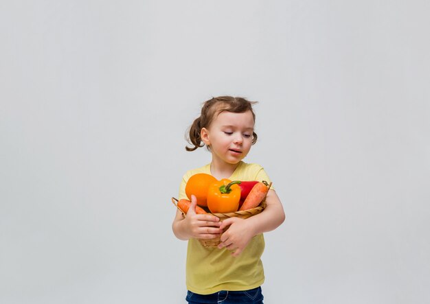 A little girl holds a basket of vegetables and fruit on a white space. A cute girl with pigtails look away at vegetables and fruits. Copy space.