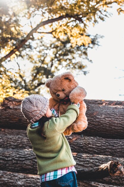 Little girl holds a bear in her arms on the background of logs on an autumn day.