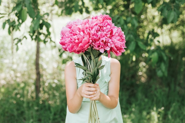 Little girl holds a bouquet of pink peony flowers in bloom in his hands and puts it over her face