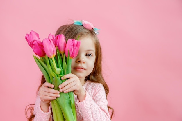 A little girl holds a bouquet of pink tulips on a pink background