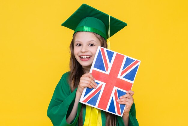 A little girl holds an english language book with the flag of great britain a student in a master's hat rejoices at graduation language school for children yellow isolated background