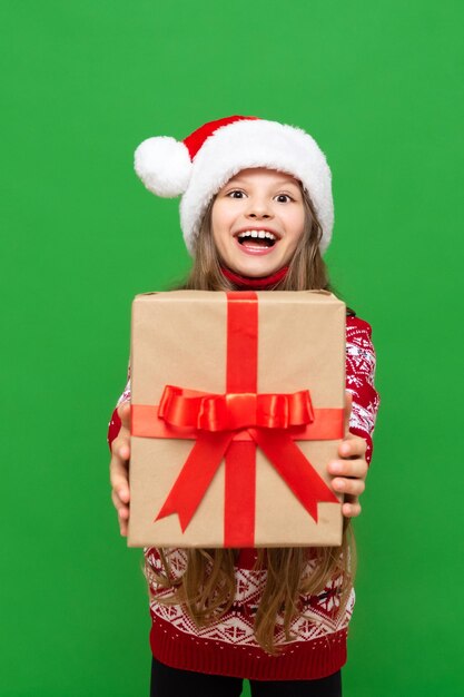 A little girl holds a gift and holds it forward on a green isolated background A child in a Santa Claus hat and a warm sweater holds a gift box