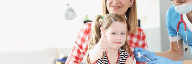 Little girl holds her thumbs up and doctor vaccinates her shoulder in medina office