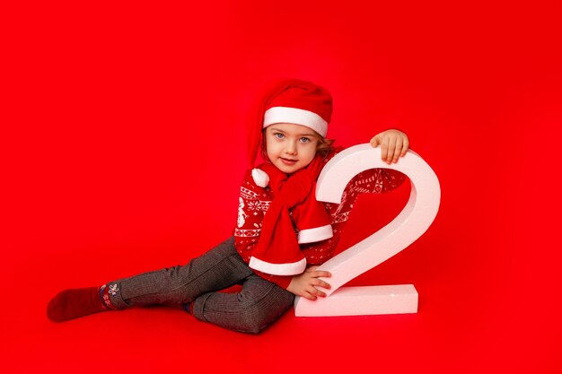 A little girl holds the number two in Santa's hat on a red background, space for text