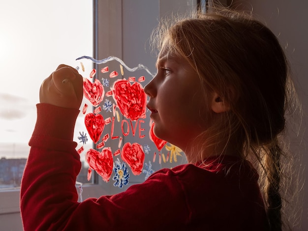 小さな女の子は絵筆を手に窓ガラスに赤いハートを描きますバレンタインデーの愛のデート検疫レジャー