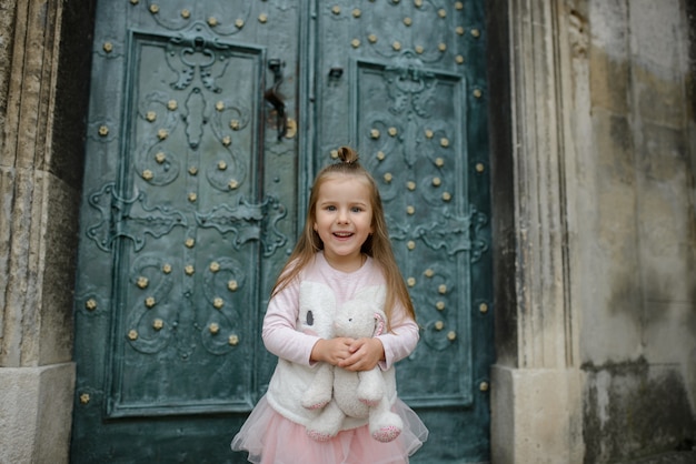 Little girl holds a toy bunny in her hands and smiles. Close-up. Girl posing on the background of the doors of the old church.