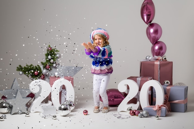 little girl in holiday decor on light , gift boxes, big numbers 2020, new year and Christmas 