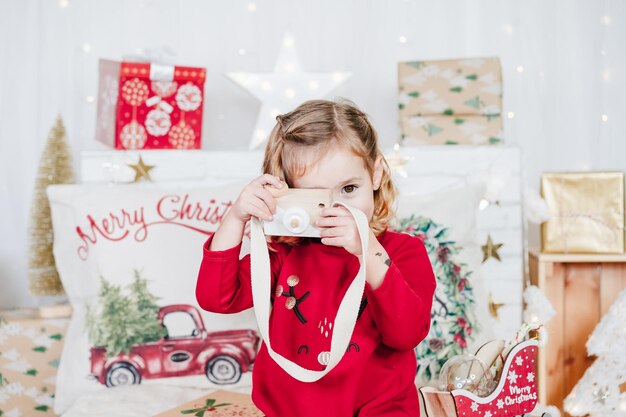 赤いクリスマスドレスを着たおもちゃのカメラを持った家にいる小さな女の子がクリスマスデコを越えて家にいる