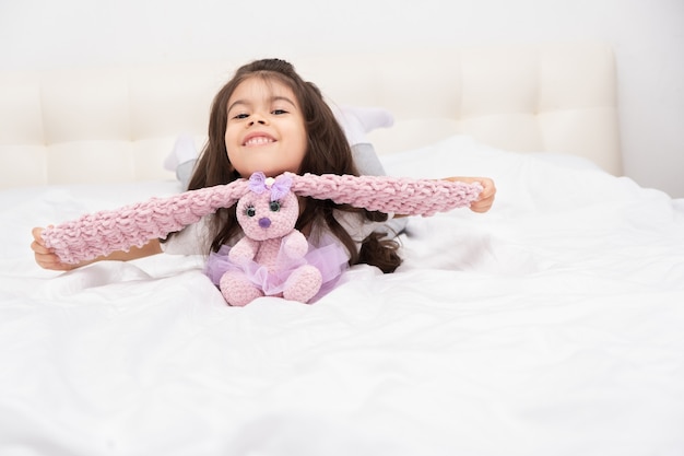 La bambina in abbigliamento domestico si siede con il coniglietto dell'orsacchiotto sul letto a casa.