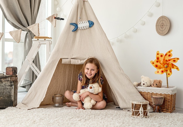 Little girl hugging plush teddy sitting in wigwam in children's room