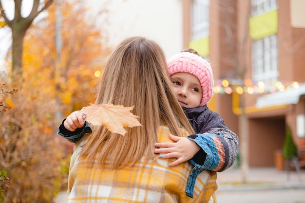 小さな女の子は、秋の通りの秋の色に沿って散歩で彼女のお母さんを抱きしめます