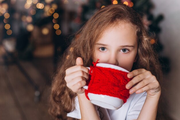 Маленькая девочка в рождественские украшения с чаем в уютном доме с красочными новогодними огнями