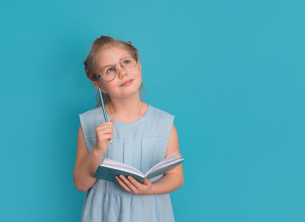Фото Маленькая девочка в очках с блокнотом или дневником и карандашом на синем фоне