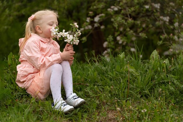 写真 庭でピンクのドレスを着た小さな女の子花がく枝と一緒に座っている女の子そして彼らの春の日を嗅いでいます