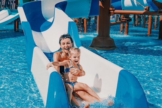 Фото Маленькая девочка в купальнике спускается по синим горкам к бассейну, мама и дочь играют и плавают в открытом бассейне
