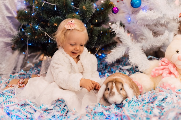 흰 드레스를 입은 어린 소녀가 토끼와 함께 크리스마스 트리 근처에서 놀고 있다