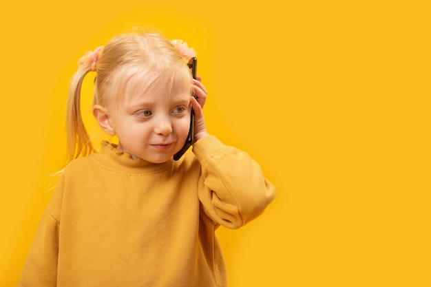 写真 電話で話す黄色いジャケットを着た小さな女の子 子供たちがスマートフォンを使用する 黄色の背景にスタジオポートレート コピースペース