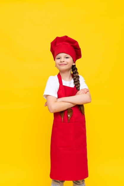 A little girl is a cook in a red apron and a cap on a yellow isolated background a girl is studying to become a cook