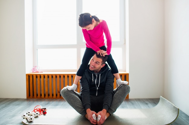 Little girl is doing stretching exercises workout at home. Cute kid and daddy are training on a mat indoor. Little dark-haired female model in sportswear has exercises near the window in her room