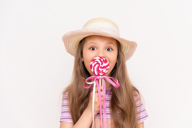 A little girl is eating a lollipop on a white isolated background A child eats sweets in a straw summer hat