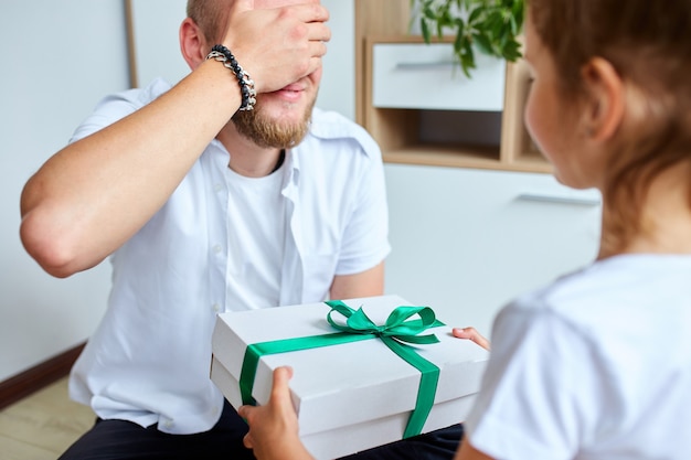 Little girl is giving her handsome father a gift box on Father's day, smiling daughter congratulating dad and giving present on birthday at home. I love you, dad. Happy Father's Day.