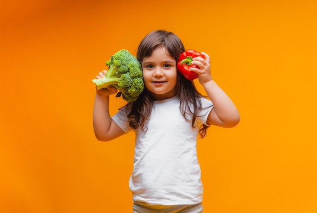 A little girl is holding a green raw broccoli and other vegetables on a yellow background