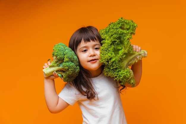 小さな女の子が黄色の背景に緑の生のブロッコリーと他の野菜を持っています