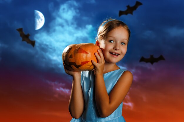 Little girl is holding a pumpkin against the background of the evening moon sky.