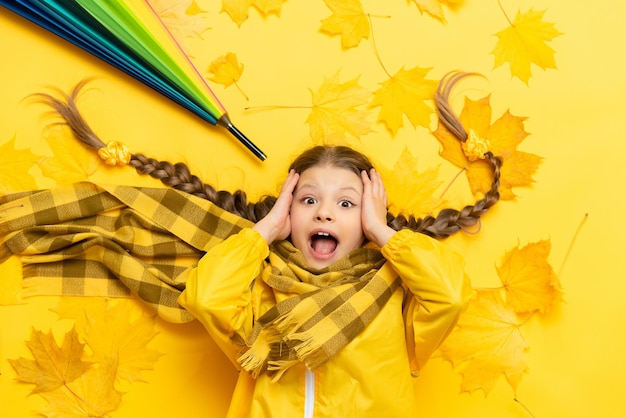 A little girl is lying on autumn maple leaves on a yellow background A child with pigtails in a raincoat and with an umbrella clutched his head with his mouth open