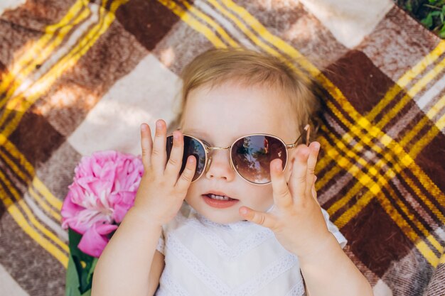 A little girl is lying on a checkered bedspread and trying on sunglasses.