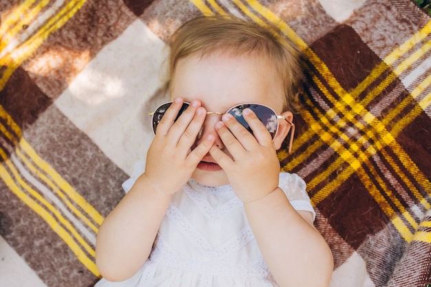 A little girl is lying on a checkered bedspread and trying on sunglasses.