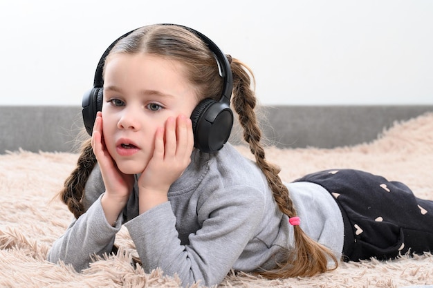 A little girl is lying on the sofa and listening to music in headphones