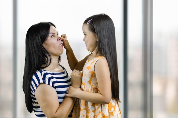 Little girl is making pig nose to her mother having fun and fooling around indoor blurred window bac