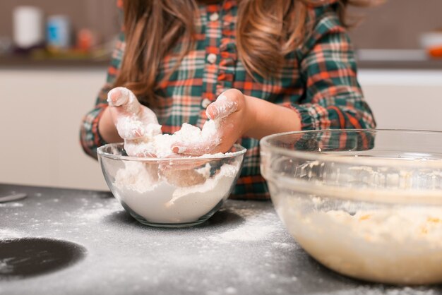 小さな女の子が家で生地用の小麦粉をボウルからボウルに注いでいます。