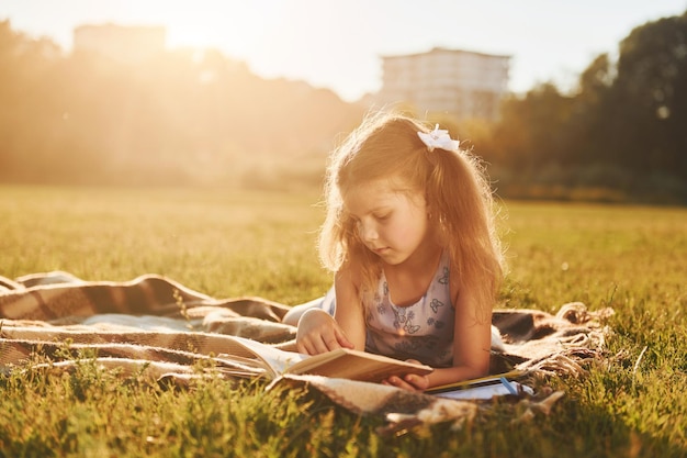 Маленькая девочка читает книгу, лежа на траве поля на открытом воздухе
