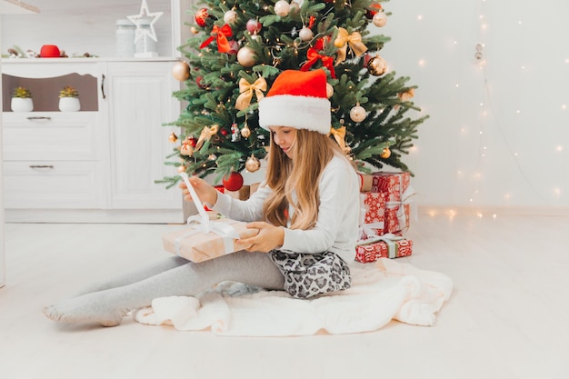 A little girl is sitting by the Christmas tree with a Christmas present. Merry Christmas.